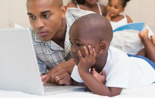 father and son looking at the computer while mother and daughter read a book in the background